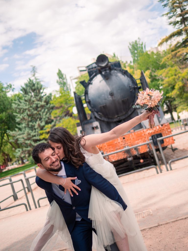 Fotografías de boda realizada por el fotógrafo de bodas Dani Pardo Studio. Una pareja de novios salen en una escena, donde la novia está subida a la espalda del novio dándole un beso en la mejilla. Detrás aparece el frontal de un tren.
