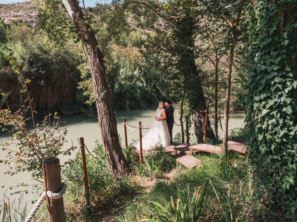 Fotografías de boda realizada por el fotógrafo de bodas Dani Pardo Studio. Una pareja de novios posa en una estructura de madera entre árboles junto a un río.